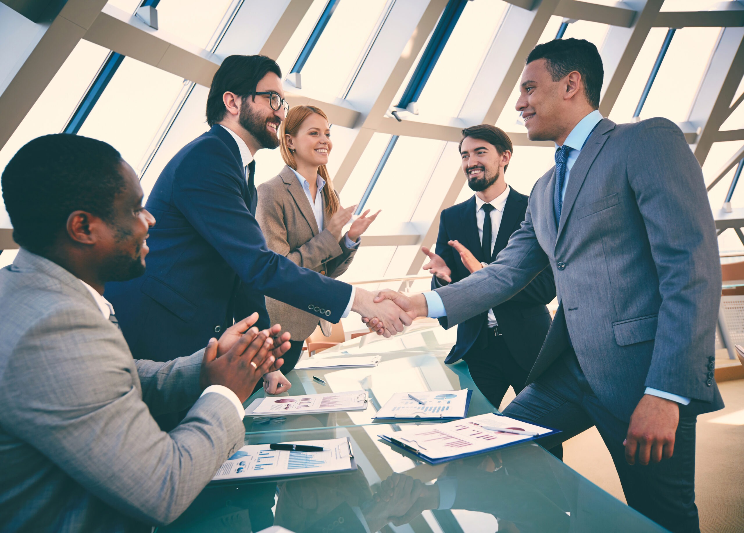 Business professionals shake hands in a modern office, celebrating a successful agreement. Team members applaud, showcasing collaboration and success.